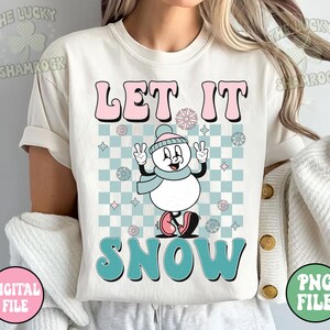 May include: Cream-colored t-shirt with the words "LET IT SNOW" in pink and teal bubble letters. A cartoon snowman with a blue scarf and hat is in the center, set against a checkered background. The shirt has rolled-up sleeves.
