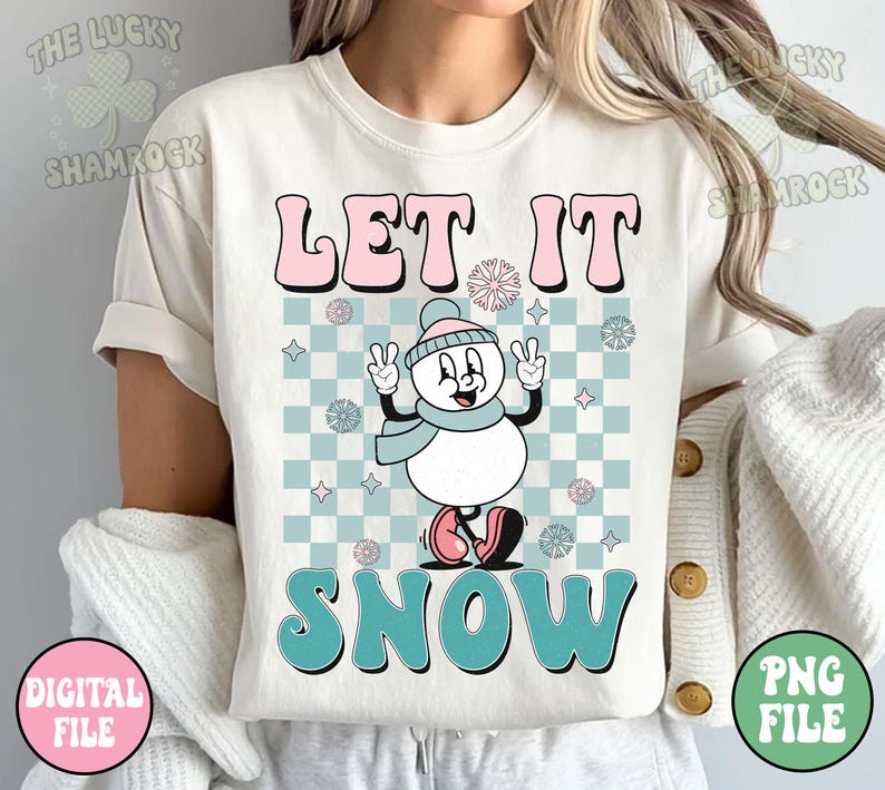May include: Cream-colored t-shirt with the words "LET IT SNOW" in pink and teal bubble letters. A cartoon snowman with a blue scarf and hat is in the center, set against a checkered background. The shirt has rolled-up sleeves.