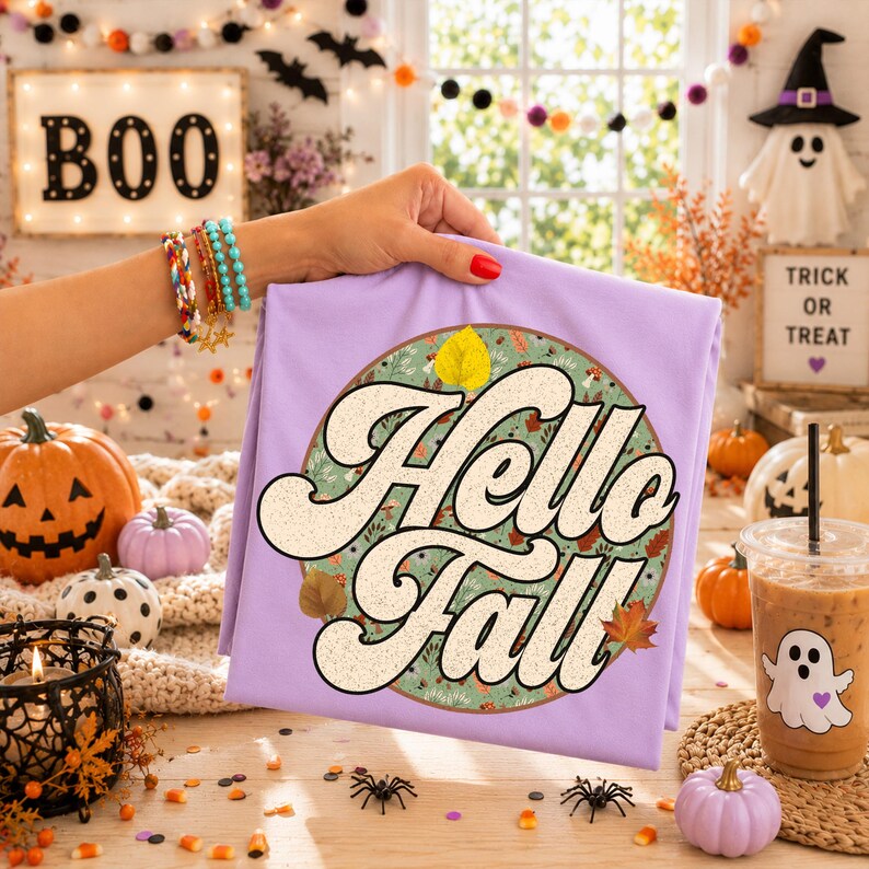 May include: A light purple t-shirt featuring a circular graphic with a green background, autumn leaves, and the text Hello Fall in large, cream-colored retro-style lettering. The design is centered on the front of the folded garment.