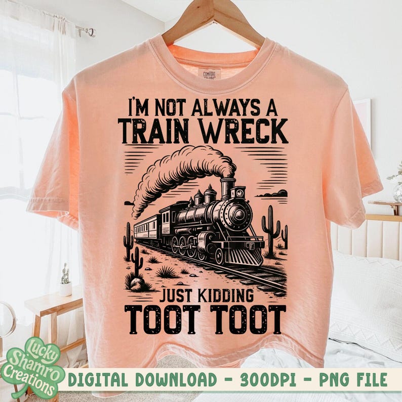 K&ouml;nnte beinhalten: Pfirsichfarbenes T-Shirt mit einer schwarzen Grafik einer alten Dampflokomotive, aus deren Schornstein Rauch aufsteigt. Der Text lautet "I'M NOT ALWAYS A TRAIN WRECK JUST KIDDING TOOT TOOT". Das T-Shirt h&auml;ngt an einem Holzb&uuml;gel.