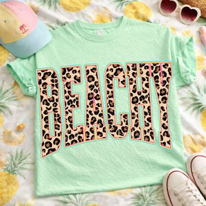 May include: A light green t-shirt with the word "BEACHY" in large, bold letters. The letters have a leopard print design with a light blue outline. The shirt is made of a soft material and is perfect for a day at the beach.