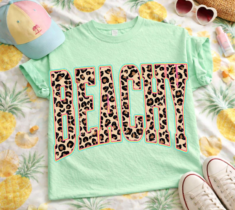 May include: A light green t-shirt with the word "BEACHY" in large, bold letters. The letters have a leopard print design with a light blue outline. The shirt is made of a soft material and is perfect for a day at the beach.