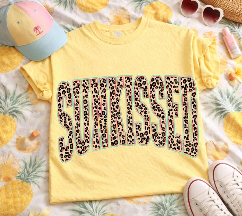 May include: A bright yellow short-sleeved t-shirt featuring the word SUNKISSED printed in a large, bold, arched font. The letters have a brown and black leopard print pattern with a thin teal outline. The shirt is made of a solid yellow fabric.