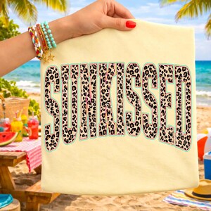May include: A light yellow t-shirt with the word "SUNKISSED" in large, leopard print letters. The letters have a light green outline. The background shows a beach scene with a picnic table, drinks, and the ocean.