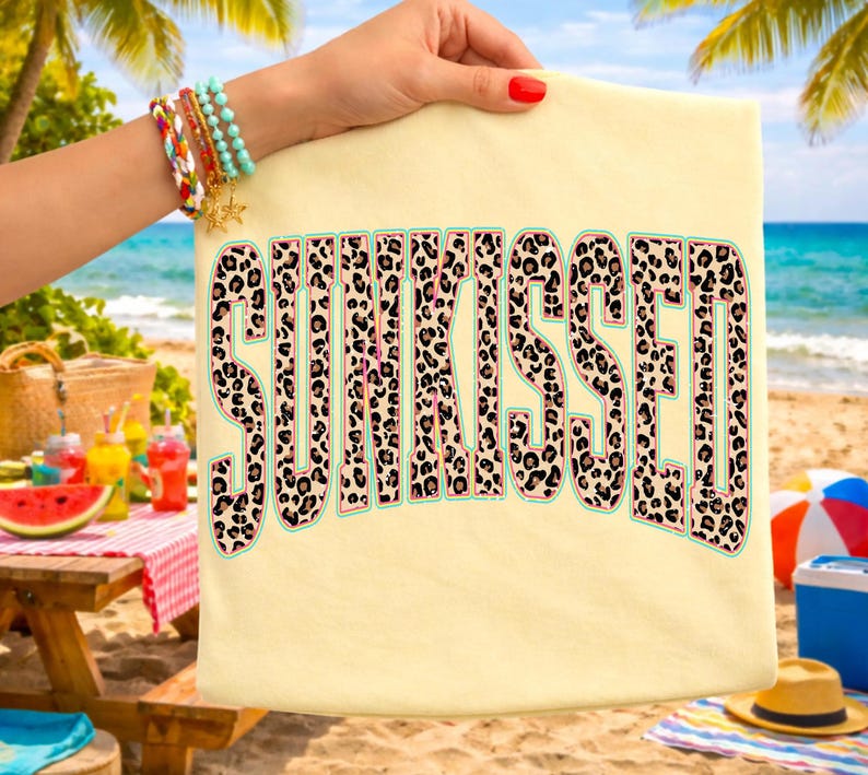 May include: A light yellow t-shirt with the word "SUNKISSED" in large, leopard print letters. The letters have a light green outline. The background shows a beach scene with a picnic table, drinks, and the ocean.