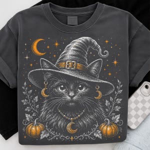 May include: A charcoal gray t-shirt featuring a graphic illustration of a black cat wearing a witch hat, gold crescent moon earrings, and a matching crescent moon necklace. The design includes orange pumpkins, stars, and white leaf accents.