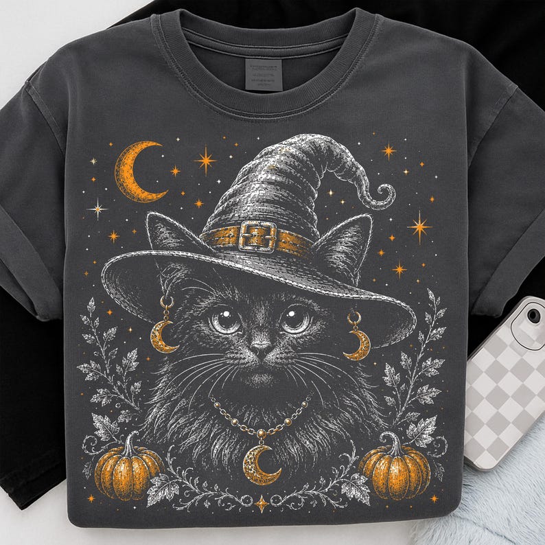 May include: A charcoal gray t-shirt featuring a graphic illustration of a black cat wearing a witch hat, gold crescent moon earrings, and a matching crescent moon necklace. The design includes orange pumpkins, stars, and white leaf accents.