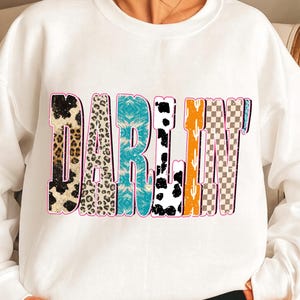 May include: White sweatshirt with the word "DARLIN'" in a colorful, patterned design. The letters feature animal print, turquoise, black and white cow spots, orange, cactus, and checkered patterns, outlined in pink.