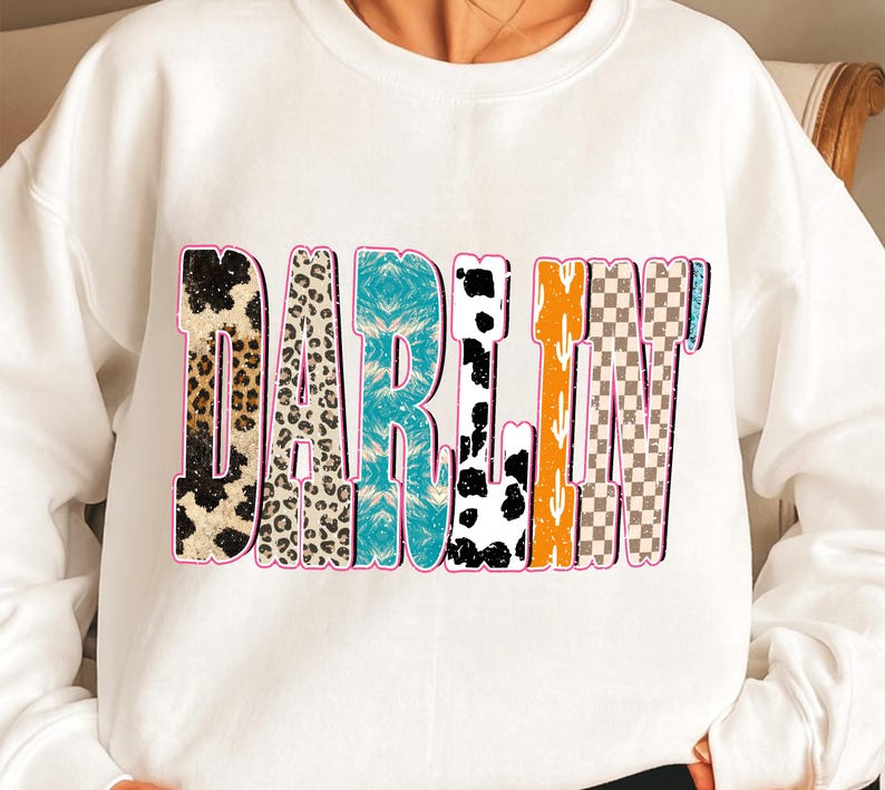 May include: White sweatshirt with the word "DARLIN'" in a colorful, patterned design. The letters feature animal print, turquoise, black and white cow spots, orange, cactus, and checkered patterns, outlined in pink.