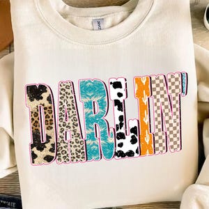 May include: Cream-colored sweatshirt with the word "DARLIN" in a colorful, patterned design. The letters feature animal prints, turquoise, cow print, orange cactus, and a checkered pattern, outlined in pink.