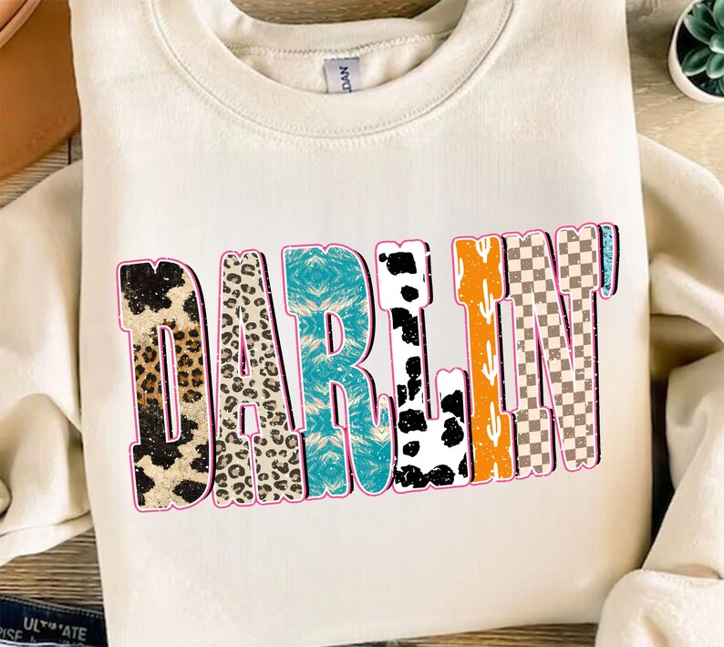 May include: Cream-colored sweatshirt with the word "DARLIN" in a colorful, patterned design. The letters feature animal prints, turquoise, cow print, orange cactus, and a checkered pattern, outlined in pink.