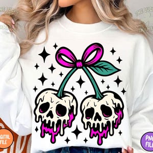 May include: White sweatshirt featuring a graphic of two cherries with skull faces, dripping with pink liquid. The cherries are connected by green stems and a pink bow. Black star shapes surround the cherries.