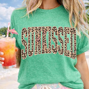 May include: A heather green short-sleeve t-shirt featuring the word SUNKISSED printed in a large, bold, arched font. The letters have a brown and black leopard print pattern with a thin light pink outline against the green fabric background.