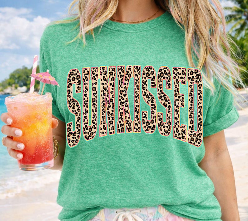 May include: A heather green short-sleeve t-shirt featuring the word SUNKISSED printed in a large, bold, arched font. The letters have a brown and black leopard print pattern with a thin light pink outline against the green fabric background.