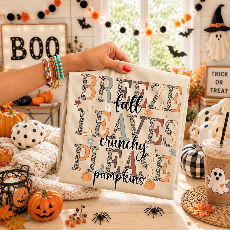 May include: A beige t-shirt featuring the text BREEZE FALL LEAVES crunchy PLEASE pumpkins in a mix of leopard print and solid muted colors. The design includes small star and flower accents, with the words arranged in a stacked, block-letter style.