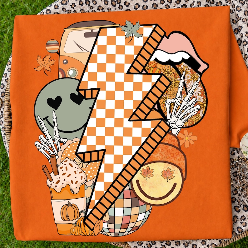 May include: An orange t-shirt featuring a large central white and orange checkered lightning bolt graphic. The design includes a green smiley face with heart eyes, a skeleton hand, a pumpkin spice drink, a disco ball, and autumn leaves.