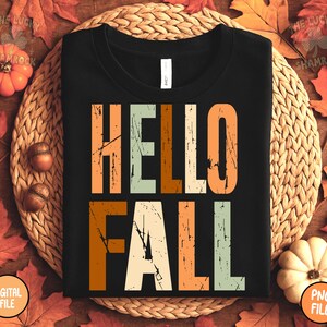 May include: Black t-shirt with the words "HELLO FALL" in a distressed, multi-colored font. The text is in shades of orange, beige, and green. The shirt is displayed on a woven mat with fall leaves and pumpkins.
