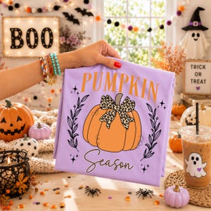 May include: A light purple t-shirt featuring a large orange pumpkin graphic with a leopard print bow. The text "PUMPKIN" is printed in orange above the pumpkin, and the word "Season" is written in black cursive script below it.