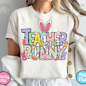 以下が含まれることがあります： 「Teacher Bunny」と書いてあるカラフルなグラフィックデザインが施された白いTシャツ。グラフィックには、ピンクの耳をしたウサギ、イースターエッグ、その他のイースターをテーマにした要素が描かれています。