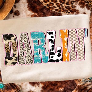May include: Cream-colored t-shirt with the word "DARLIN" in a multi-patterned design. The letters feature leopard print, turquoise, cow print, orange cactus, and a checkered pattern, outlined in pink.