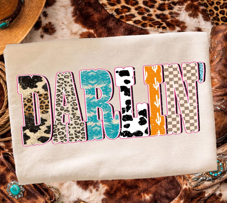 May include: Cream-colored t-shirt with the word "DARLIN" in a multi-patterned design. The letters feature leopard print, turquoise, cow print, orange cactus, and a checkered pattern, outlined in pink.