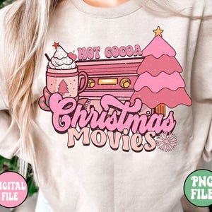 K&ouml;nnte beinhalten: Ein beiger Pullover mit einem rosafarbenen und wei&szlig;en Grafikdesign, das eine Tasse hei&szlig;e Schokolade, einen Weihnachtsbaum und einen Vintage-Kassettenrekorder mit dem Text "Hot Cocoa Christmas Movies" zeigt.