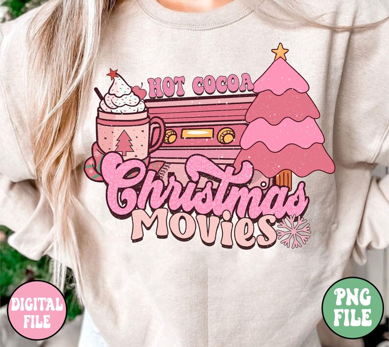 K&ouml;nnte beinhalten: Ein beiger Pullover mit einem rosafarbenen und wei&szlig;en Grafikdesign, das eine Tasse hei&szlig;e Schokolade, einen Weihnachtsbaum und einen Vintage-Kassettenrekorder mit dem Text "Hot Cocoa Christmas Movies" zeigt.