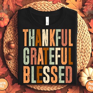 May include: Black t-shirt with the words "THANKFUL, GRATEFUL, BLESSED" in a distressed, vintage-style font. The text is in shades of orange, brown, and green. The shirt is displayed on a woven mat with fall leaves and pumpkins.