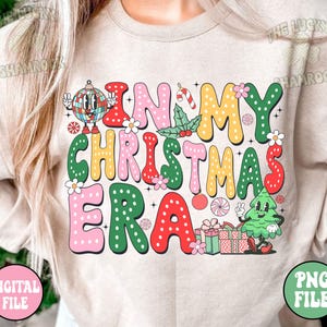 May include: Beige sweatshirt with the phrase "IN MY CHRISTMAS ERA" in colorful, retro-style lettering. The design includes a disco ball, candy cane, holly, and a cartoon Christmas tree. The words are outlined in various colors.