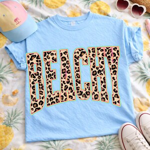 May include: Light blue t-shirt with the word "BEACHY" in a leopard print design. The word is outlined in a teal color with a pink inner border. A light blue baseball cap and white sneakers are also in the image.