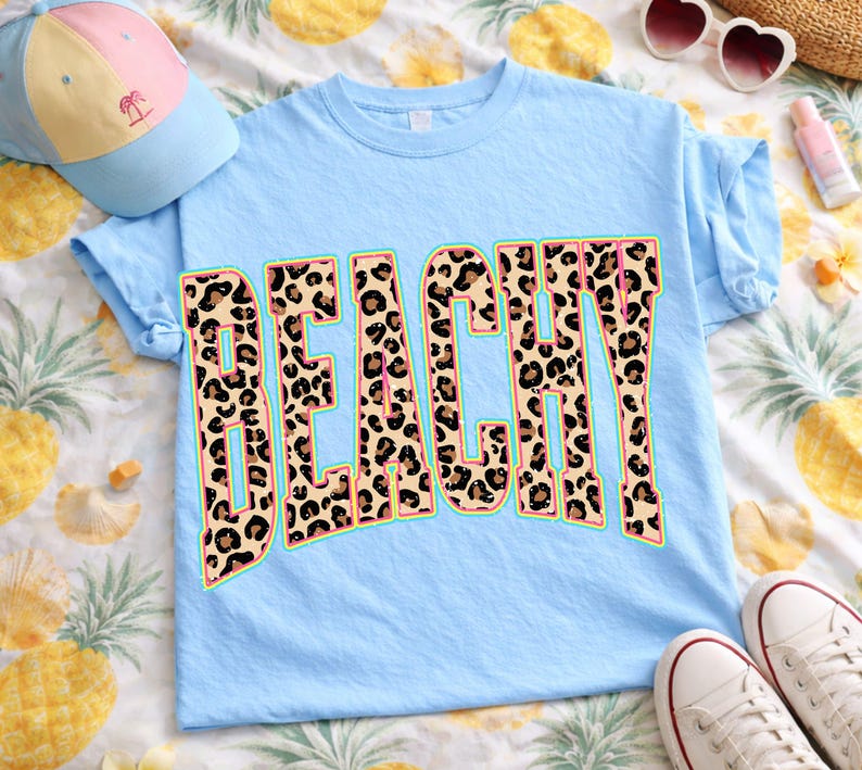 May include: Light blue t-shirt with the word "BEACHY" in a leopard print design. The word is outlined in a teal color with a pink inner border. A light blue baseball cap and white sneakers are also in the image.