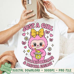 May include: White t-shirt featuring a cartoon character with a pink body, a yellow bow, and a drink. The text "JUST A GIRL WITH BIG DREAMS & MATCHA ENERGY" is displayed in pink. Additional graphics include hearts, flowers, and bows. Digital download.