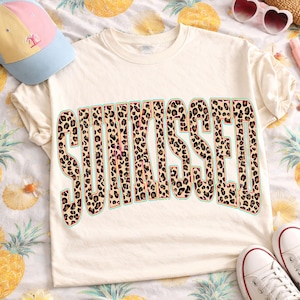 May include: A cream-colored short-sleeve t-shirt featuring the word SUNKISSED in large, block-style letters with a brown and black leopard print pattern. The text is outlined in a thin teal border and centered on the front of the garment.