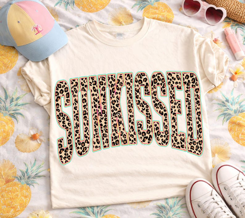 May include: A cream-colored short-sleeve t-shirt featuring the word SUNKISSED in large, block-style letters with a brown and black leopard print pattern. The text is outlined in a thin teal border and centered on the front of the garment.