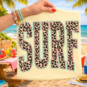 May include: A light yellow t-shirt with the word "SURF" in large, bold letters. The letters are filled with a leopard print pattern, outlined in pink and turquoise. The shirt is held up by a hand with bracelets, with a beach scene in the background.