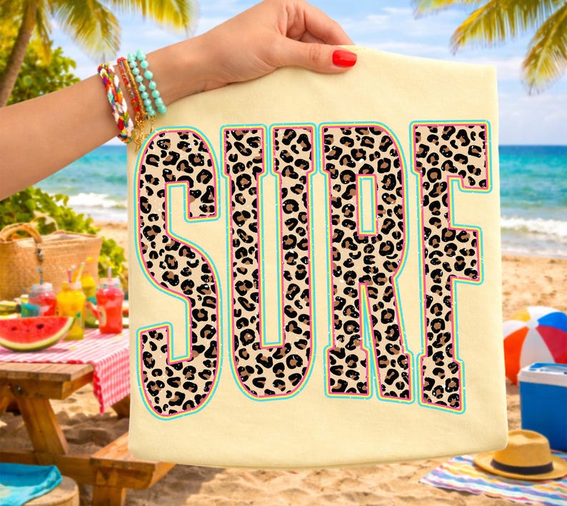 May include: A light yellow t-shirt with the word "SURF" in large, bold letters. The letters are filled with a leopard print pattern, outlined in pink and turquoise. The shirt is held up by a hand with bracelets, with a beach scene in the background.