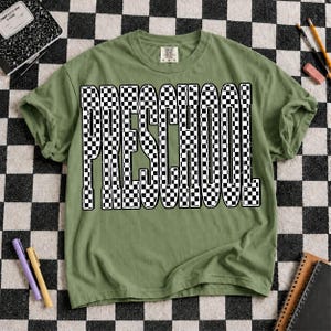 K&ouml;nnte beinhalten: Ein olivgr&uuml;nes Kurzarm-T-Shirt mit dem Wort "PRESCHOOL", das in einem auff&auml;lligen schwarz-wei&szlig;en Schachbrettmuster gedruckt ist. Die Buchstaben haben ein abgenutztes, strukturiertes Aussehen. Das T-Shirt hat einen Rundhalsausschnitt.