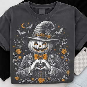 May include: A charcoal gray t-shirt featuring a graphic of a pumpkin-headed figure wearing a witch hat and a suit with a bow tie. The figure is making a heart shape with its hands, surrounded by orange maple leaves, bats, and stars.