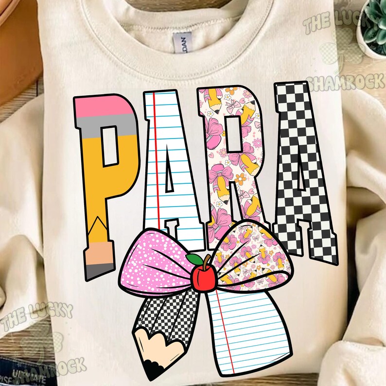 K&ouml;nnte beinhalten: Ein cremefarbenes Sweatshirt mit Rundhalsausschnitt und dem Wort PARA in gro&szlig;en Blockbuchstaben. Die Buchstaben sind mit Mustern aus Bleistiften, linierten Bl&auml;ttern, Blumen-Bleistiften und schwarz-wei&szlig;em Schachbrettmuster gef&uuml;llt, erg&auml;nzt durch eine gro&szlig;e rosa Schleife und einen Apfel.