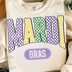 May include: Cream-coloured sweatshirt featuring the words "MARDI GRAS" in large, colourful, checkered letters. "MARDI" is in purple, yellow, and green, while "GRAS" is in a purple oval. A casual garment.