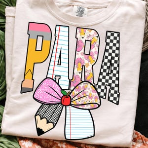 K&ouml;nnte beinhalten: Ein hellbeiges T-Shirt mit dem Wort PARA in gro&szlig;en Blockbuchstaben. Die Buchstaben sind mit Bleistift-, linierten Papier-, Blumen- und Schachbrettmustern gef&uuml;llt. Unter dem Text befindet sich eine gro&szlig;e Schleifengrafik mit einem roten Apfel.