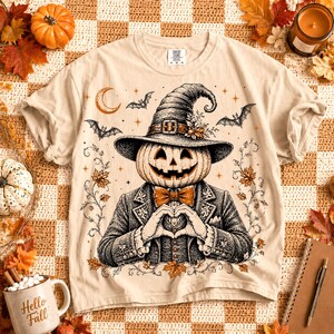 May include: A beige t-shirt featuring a black and white graphic of a pumpkin-headed figure wearing a witch hat and a suit with a bow tie. The figure is making a heart shape with their hands. The design includes bats, a crescent moon, and leaves.