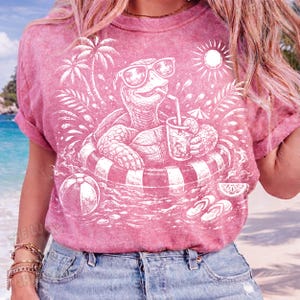 May include: A pink t-shirt featuring a white graphic of a turtle wearing sunglasses and floating in a striped inner tube, drinking a beverage with a straw. The design includes a beach ball, palm trees, and a sun.