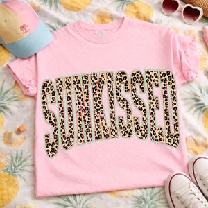 May include: A light pink short-sleeve t-shirt featuring the word SUNKISSED printed in a large, bold, arched font. The letters have a brown and black leopard print pattern with a thin teal outline. The shirt has a crew neckline and short sleeves.