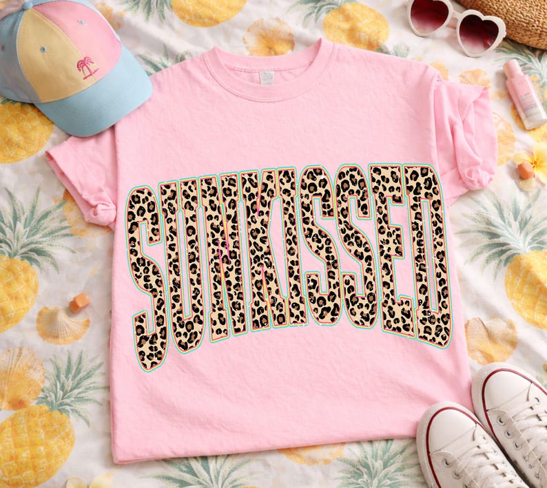 May include: A light pink short-sleeve t-shirt featuring the word SUNKISSED printed in a large, bold, arched font. The letters have a brown and black leopard print pattern with a thin teal outline. The shirt has a crew neckline and short sleeves.