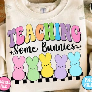 以下が含まれることがあります： 虹色の文字で「TEACHING Some Bunnies」と書かれた白いスウェットシャツ。文字は市松模様と5つのパステルカラーのウサギのシルエットで囲まれています。
