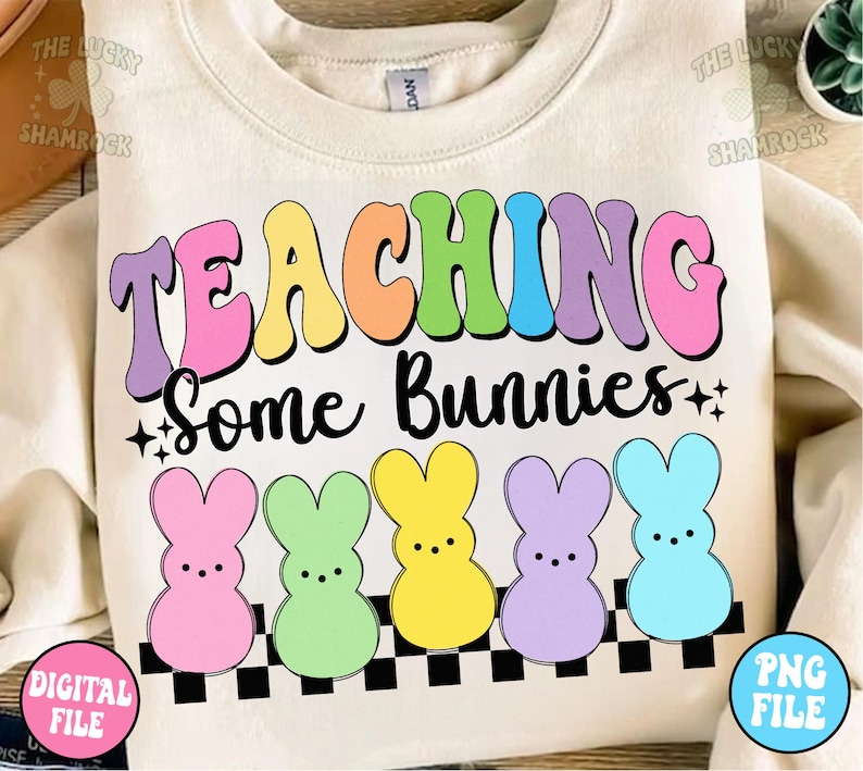 以下が含まれることがあります： 虹色の文字で「TEACHING Some Bunnies」と書かれた白いスウェットシャツ。文字は市松模様と5つのパステルカラーのウサギのシルエットで囲まれています。
