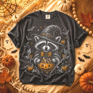 May include: A charcoal gray t-shirt featuring a graphic of a raccoon wearing a witch hat and a polka-dot bow tie, holding a jack-o'-lantern filled with candy corn. The design includes stars, bats, and swirling vines in white and orange.