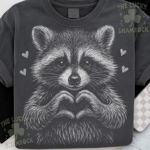 Peut inclure: T-shirt gris fonc&eacute; avec une illustration blanche d&eacute;taill&eacute;e d'un raton laveur formant un c&oelig;ur avec ses mains. Le motif comprend de petits c&oelig;urs blancs et le texte "THE LUCKY SHAMROCK".