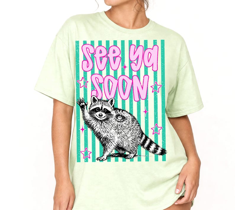 May include: A pale green t-shirt with a graphic of a waving raccoon and the words "See Ya Soon" in pink bubble letters. The raccoon is black and white, set against a teal and white striped background with pink stars.